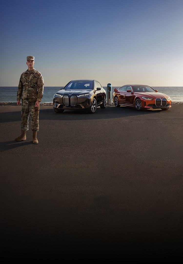 A BMW iX xDrive50 and BMW i4 eDrive35 in front a beach landscape with a U.S. Military member posed in the front.